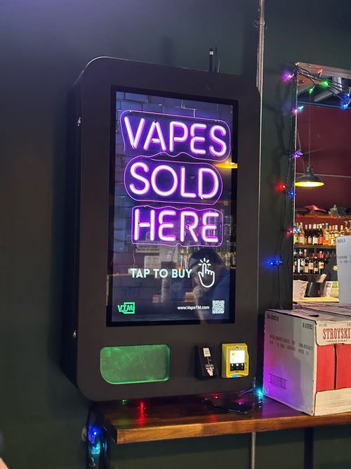 Vape vending machine mounted beside a bar, showing a neon Vapes Sold Here screen with a tap to buy prompt